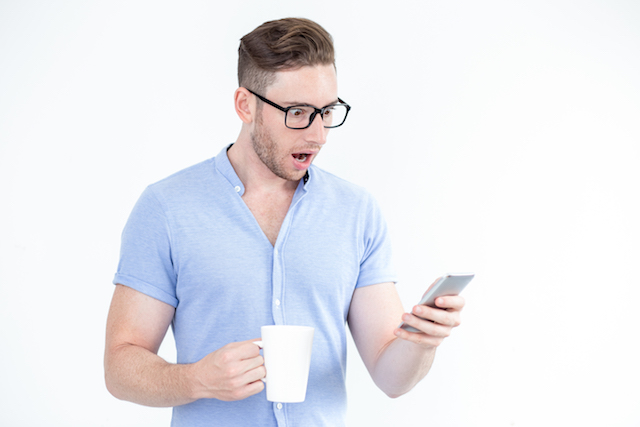 Man holding coffee cup while scrolling instagram on phone, looking shocked.