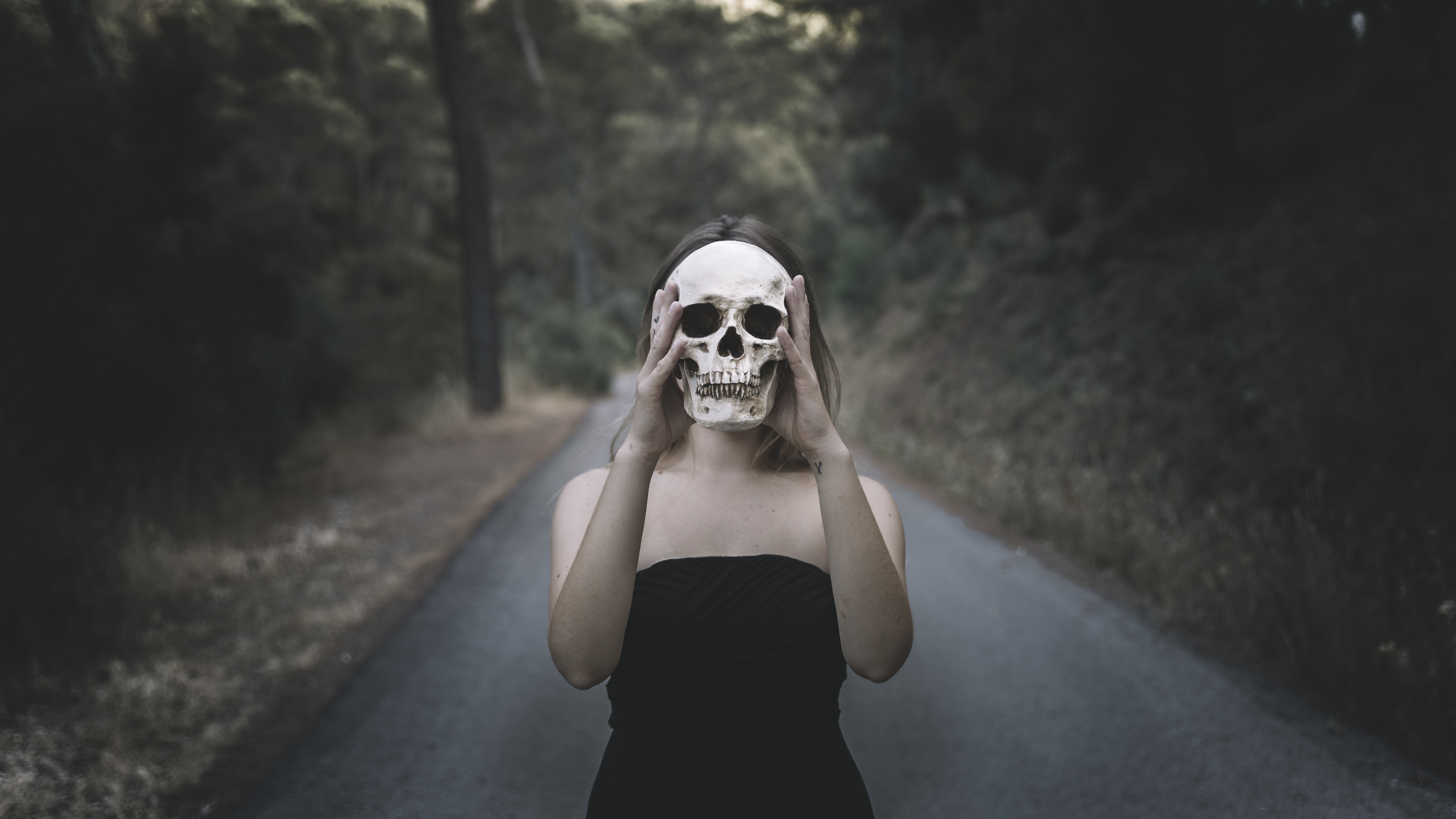 woman holds a skull in front of her face while she stands on a dark forested road. Elements of nightmares.